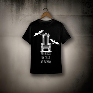 May include: Black t-shirt with a white graphic of a throne with two bats flying above it. The text "MY HOUSE. MY CHAIR. MY WOMAN." is printed below the throne.