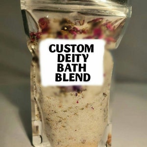 May include: A clear plastic bag filled with a white bath salt blend with dried flowers and herbs. The bag has a label that reads "CUSTOM DEITY BATH BLEND". A small wooden scoop is next to the bag.