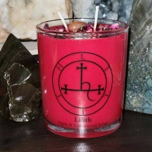 May include: A red candle with a black sigil of Lilith, the demoness, on the front. The candle is labeled "Lilith Apple & Sandalwood Scent with Carnelian, Rose & Patchouli".