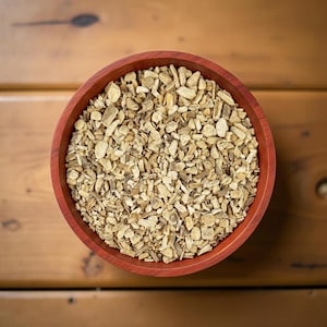 May include: A top-down view of a round, red wooden bowl filled with chopped, beige-colored herbs. The bowl sits on a wooden surface with a warm, brown tone. The herbs appear to be dried and have a rough texture.