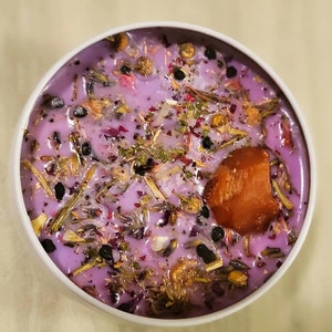 May include: A purple candle with dried flowers and herbs embedded in the wax. The candle is in a white jar with a clear lid. There is a small piece of orange wax on top of the candle.