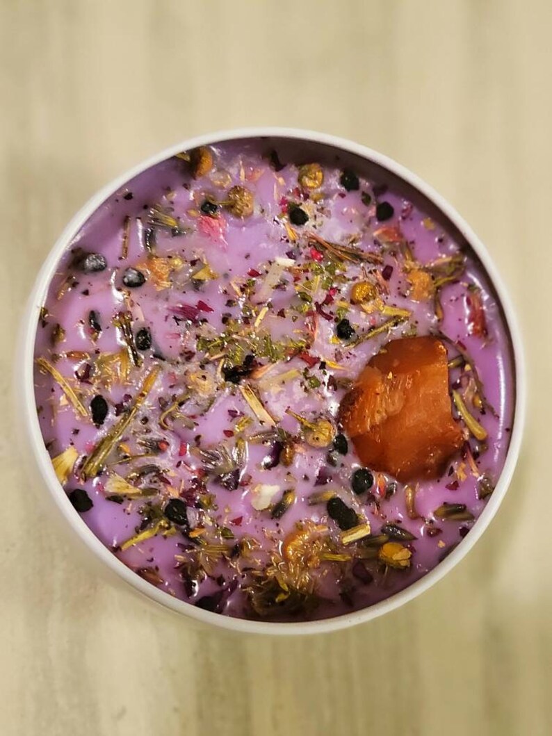 May include: A purple candle with dried flowers and herbs embedded in the wax. The candle is in a white jar with a clear lid. There is a small piece of orange wax on top of the candle.