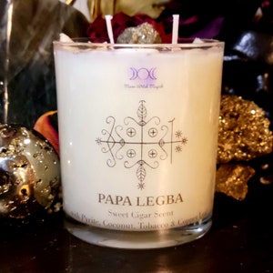 Op de afbeelding: Een witte kaars in een heldere glazen pot met twee lonten. De kaars heeft een zwart grafisch ontwerp en de woorden "PAPA LEGBA" en "Sweet Cigar Scent" erop gedrukt. De tekst "Moon Witch Magich" is ook zichtbaar. De kaars is versierd met pyriet.