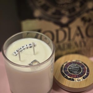 Op de afbeelding: Een witte kaars in een glazen pot met een houten deksel. De kaars heeft drie lonten en het woord "VAMPIRE" is gespeld met witte lettertegels op de was. Het deksel heeft een zwart-witte zodiac-wielontwerp met de woorden "Zodiac Candle" erop gedrukt.