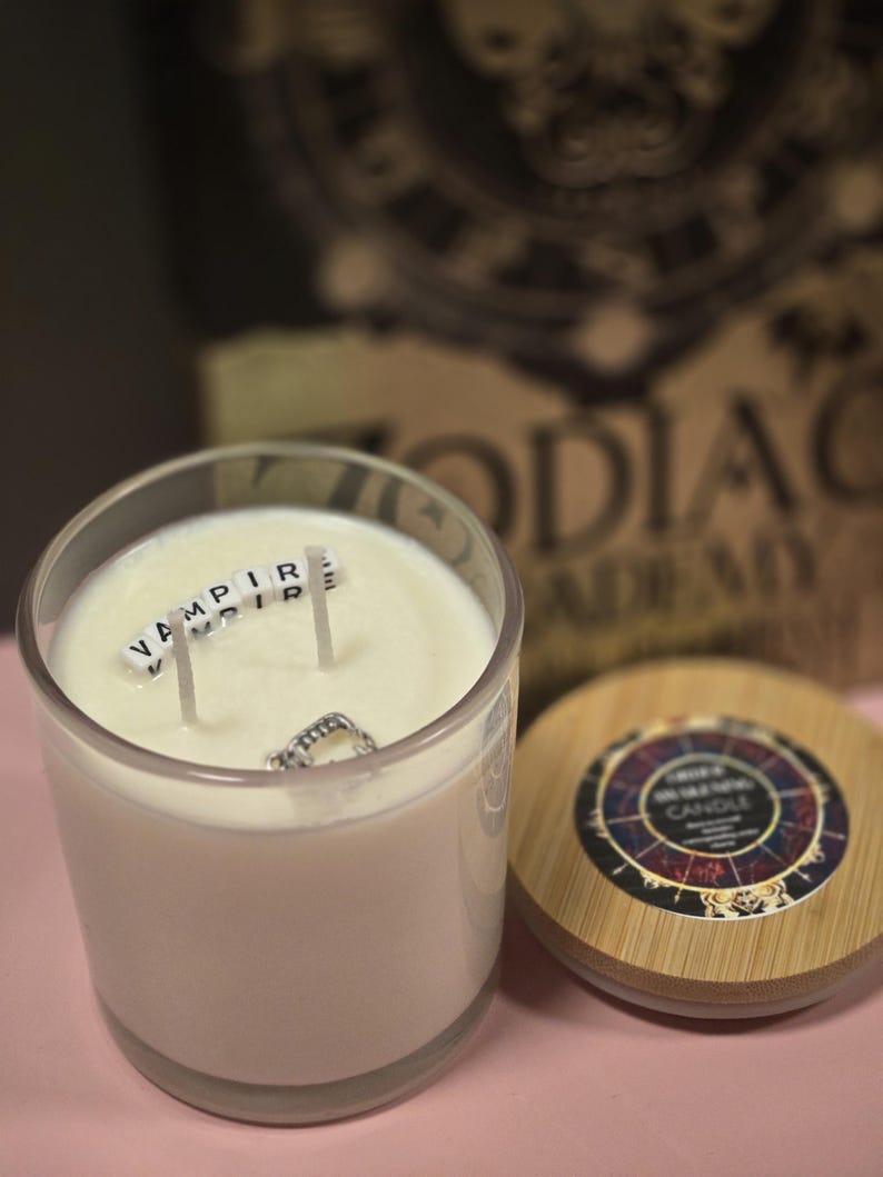 Op de afbeelding: Een witte kaars in een glazen pot met een houten deksel. De kaars heeft drie lonten en het woord "VAMPIRE" is gespeld met witte lettertegels op de was. Het deksel heeft een zwart-witte zodiac-wielontwerp met de woorden "Zodiac Candle" erop gedrukt.
