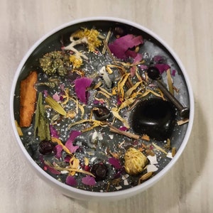 May include: A black candle with various dried herbs, flowers, and a black stone embedded in the wax. The candle is in a white container.