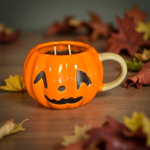 May include: A ceramic pumpkin-shaped mug with a carved jack-o'-lantern face. The orange mug has a light green handle and contains two white candles. Autumn leaves surround the mug, creating a festive fall scene.
