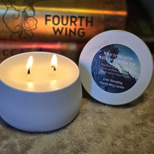 Può includere: Una candela bianca a due stoppini con fiamme. La candela è in un contenitore rotondo e bianco. Il coperchio della candela riporta il testo "WHICH DRAGON WILL YOU BOND TO?" e "Color Revealing Fourth Wing Candle."