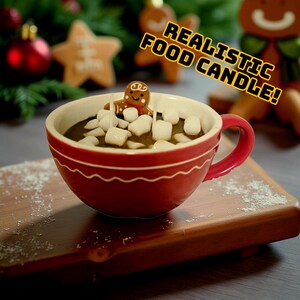 May include: A red ceramic mug filled with a dark liquid and topped with white marshmallows and a gingerbread man. The mug has a white wavy design and a red handle. The text "REALISTIC FOOD CANDLE!" is in the background.