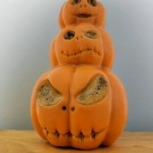 May include: A stack of three orange pumpkin-shaped candles with carved faces. The bottom pumpkin has a wide, toothy grin and large eyes. The middle pumpkin has a more neutral expression with smaller eyes. The top pumpkin has a wide smile and small eyes.