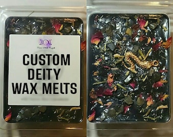Può includere: Due cere per la fusione in un imballaggio di plastica trasparente. La cera a sinistra ha un'etichetta bianca con il testo "CUSTOM DEITY WAX MELTS". La cera a destra è nera con accenti dorati, rossi e verdi.