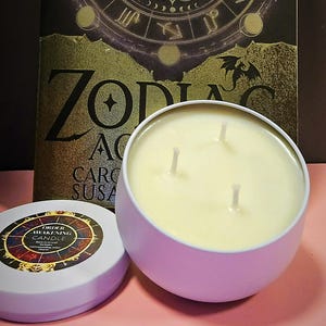 Op de afbeelding: Een witte kaars in een witte blik met drie lonten. De kaars is gelabeld "Order Awakening Candle" en heeft een ontwerp van een dierenriemwiel. De kaars staat op een roze oppervlak naast een boek met het woord "Zodiac" op de omslag.