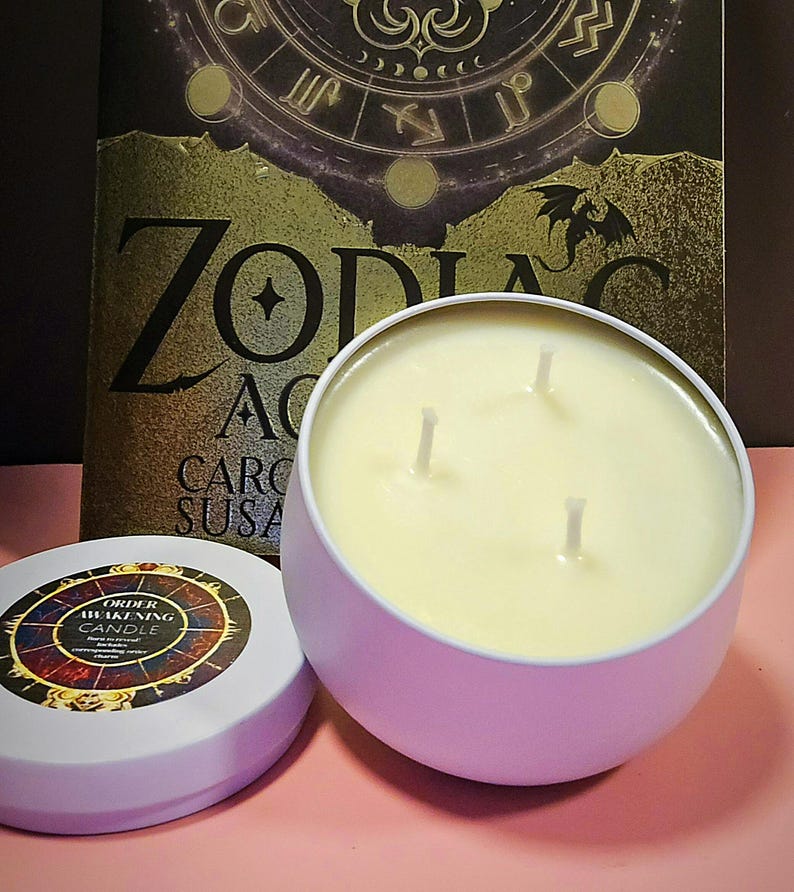 Op de afbeelding: Een witte kaars in een witte blik met drie lonten. De kaars is gelabeld "Order Awakening Candle" en heeft een ontwerp van een dierenriemwiel. De kaars staat op een roze oppervlak naast een boek met het woord "Zodiac" op de omslag.