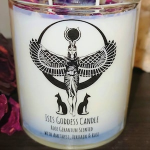 May include: A white candle with a black and white image of the Egyptian goddess Isis with wings and two cats. The candle is labeled "Isis Goddess Candle Rose Geranium Scented with Amethyst, Vervain & Rose".