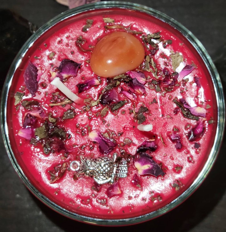 May include: A red candle with dried rose petals, herbs, and a carnelian crystal. The candle is in a clear glass jar.
