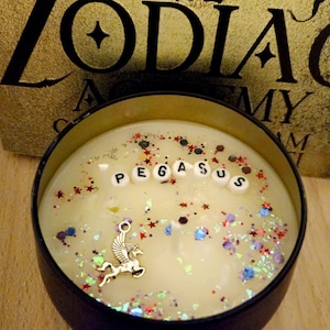 Op de afbeelding: Een zwarte kaars pot met een witte was basis en een zilveren Pegasus bedel. De pot is gevuld met kleurrijke glitter en het woord "PEGASUS" is gespeld in witte letters.