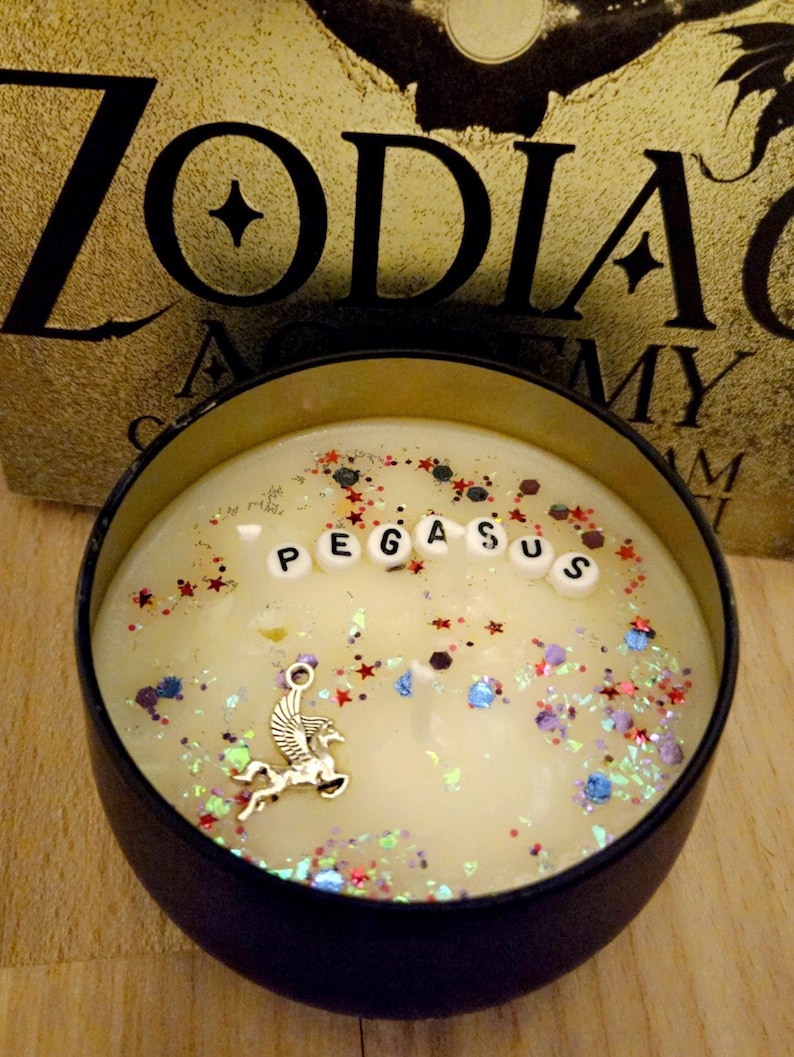 Op de afbeelding: Een zwarte kaars pot met een witte was basis en een zilveren Pegasus bedel. De pot is gevuld met kleurrijke glitter en het woord "PEGASUS" is gespeld in witte letters.
