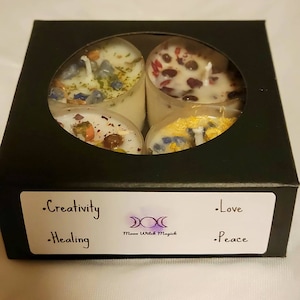 May include: A black box containing four small, white candles, each visible through oval apertures. The candles are adorned with various dried flowers and herbs. The box features a label with the words "Creativity, Healing, Love, Peace" and the logo "Moon Witch Magick".