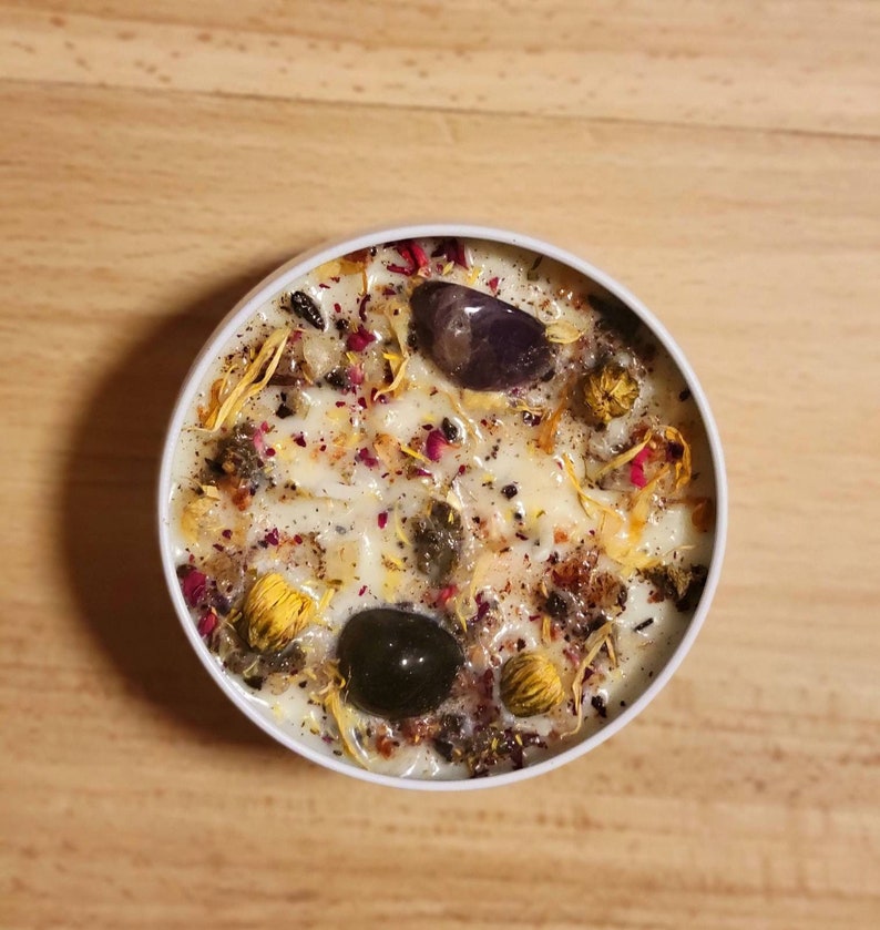 May include: A white candle with a variety of dried flowers and crystals embedded in the wax. The crystals are purple, black, and brown.