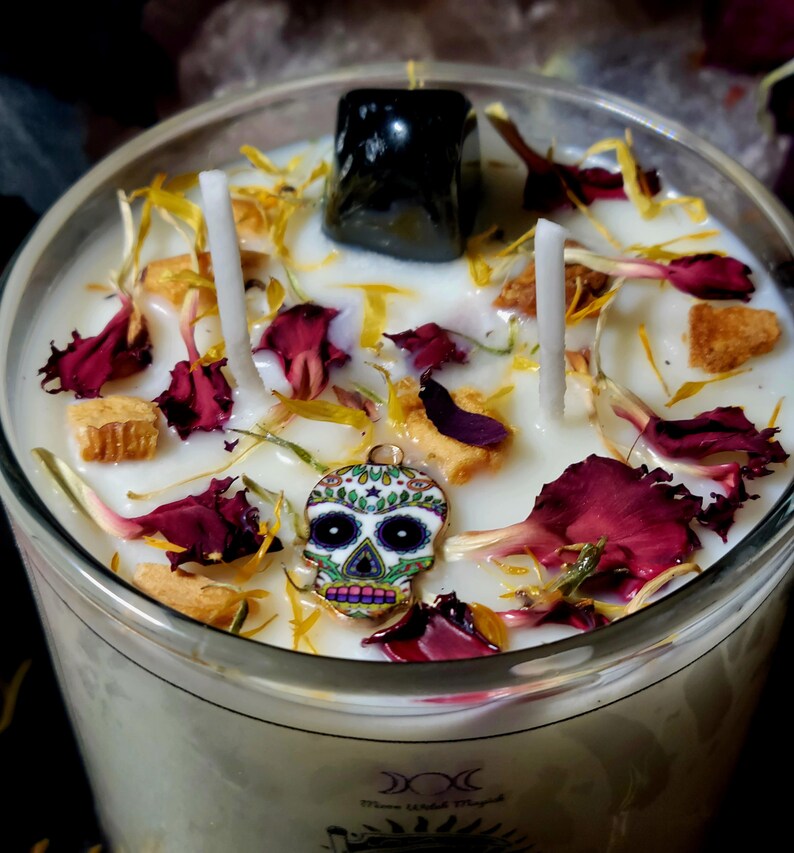 Pu&ograve; includere: Una candela bianca con un cristallo nero in cima. La candela &egrave; decorata con fiori secchi, tra cui petali gialli e garofani rossi. Un piccolo ciondolo a forma di teschio di zucchero pende dal lato della candela.