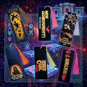 May include: A collection of black bookmarks with colorful tassel accents. Each bookmark features unique graphics and text, including phrases like "NEW ACHIEVEMENT!" and "TOO LATE! I PICKED IT." The bookmarks are set against a space-themed background.
