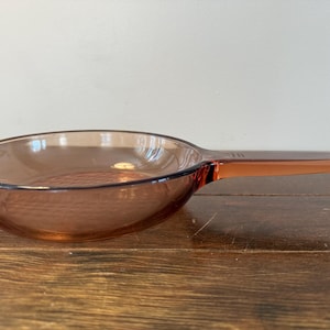 Puede incluir: Una sartén de vidrio ámbar vintage con un mango largo. La sartén tiene forma de cuenco redondeado y un fondo texturizado. El mango es una pieza sólida de vidrio, que se extiende desde el cuerpo de la sartén. La sartén está sobre una superficie de madera.