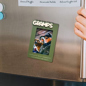 May include: A rectangular olive green magnetic photo frame featuring the word GRAMPS in raised white lettering at the top. The frame displays a photograph of a person wearing a cap and sunglasses next to a dog, with the text WORLD'S BEST GRANDPA.