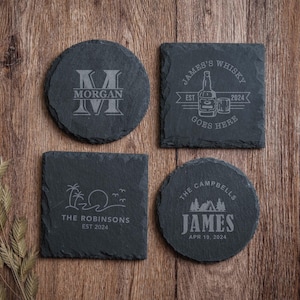 May include: Four dark gray slate coasters with rough, natural edges. Two are circular and two are square. Each features custom laser-engraved designs including the text MORGAN, JAMES'S WHISKY GOES HERE EST 2024, THE ROBINSONS EST 2024, and THE CAMPBELLS JAMES APR 19, 2024.