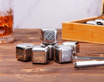 May include: A set of six polished stainless steel cubes, each featuring a laser-engraved letter Z inside a diamond-shaped border. The cubes are arranged in a cluster alongside a pair of matching metal tongs with serrated edges for gripping.