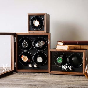 May include: Three wooden watch winder boxes with glass doors. One box holds four watches, one holds one, and one holds two. The two-watch box features the name DYLAN printed on the glass. The interiors are black with circular rotating mounts.