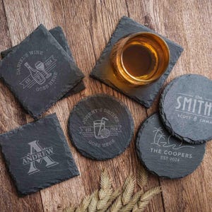 May include: A collection of personalized slate coasters with engraved designs. Square coasters feature "Daniel's Wine" and "Andrew" with a monogram. Round coasters display "Johnny's Drinks," "The Smiths," and "The Coopers" with an island scene.