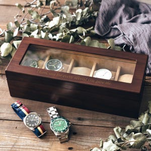 Può includere: Una scatola rettangolare per orologi in legno marrone scuro con coperchio in vetro che riporta il testo inciso "Thank you for every second you gave me." La scatola contiene cinque scomparti interni con cuscinetti color beige.