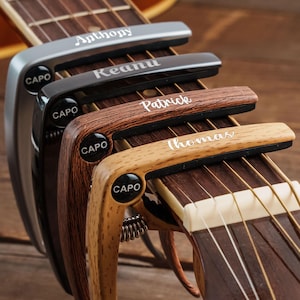 May include: Four guitar capos clamped onto a guitar neck, each featuring a unique finish and a personalized name engraved in white text. The capos are labeled with the word CAPO on the side and display the names Anthony, Keanu, Patrick, and Thomas.