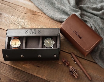 May include: A dark brown leather watch roll case with the monogram S.M.S engraved on the flap, containing two wristwatches. Beside it sits a second light brown leather watch case engraved with the name Noah, along with a wooden pen.