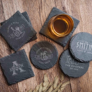 May include: A collection of personalized slate coasters with engraved designs. Square coasters feature wine and initial designs, while round coasters display drink and family name details. A glass of amber liquid sits on one coaster.