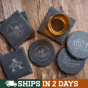 May include: A collection of square and circular dark gray slate coasters with rough, natural edges. Each coaster features custom laser-engraved text and graphics, including names like Andrew, The Smiths, and The Coopers, alongside a glass of amber liquid.