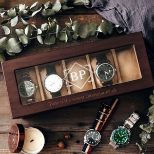 May include: A wooden watch box with a clear lid, holding four watches. The box is engraved with the initials "BP" and the quote "Time is the wisest counselor of all." Two additional watches and a candle are also visible.