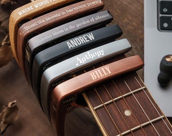 May include: A stack of six guitar capos in various finishes including wood grain, matte black, silver, and copper. Each capo is engraved with text: WHERE WORDS FAIL, MUSIC SPEAKS, Unveil the secrets in harmonic poetry, Melodies bloom in the garden of silence, ANDREW, Anthony, and BILLY.