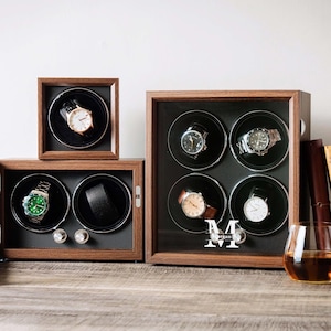 May include: Three wooden watch winders with dark brown frames and black interiors. One single-watch winder, one double-watch winder with an open door, and one quad-watch winder featuring the letter M and the name Morgan printed on the glass front.