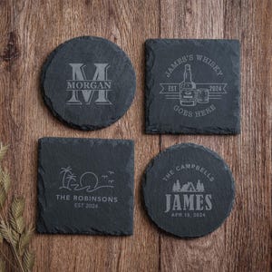 May include: Four dark gray slate coasters with rough, natural edges. Two are circular and two are square. Each features unique laser-engraved designs including a large letter M, a whiskey bottle, a beach scene with palm trees, and a cabin in the woods.