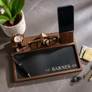 Op de afbeelding: Een houten bureau-organizer met een zwart lederen dienblad, met een horloge, bril, telefoon, pen en sleutels. De lade is gegraveerd met de naam "BARNES". Een kleine plant in een witte pot staat op de achtergrond.