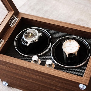 Op de afbeelding: Een houten horlogewinder met een scharnierend glazen deksel, voorzien van twee ronde roterende compartimenten bekleed met zwart materiaal. Binnenin zijn twee polshorloges op zwarte kussentjes geplaatst. Twee zilveren knoppen bevinden zich op de basis.