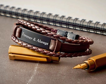 May include: A multi-strand bracelet featuring dark brown braided leather cords and a central matte black metal plate engraved with the white text Forever & Always. The bracelet includes a black metal clasp and is positioned near a gold fountain pen.