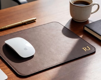May include: A rectangular brown leather mouse pad with rounded corners and stitched edges. The mouse pad features the gold embossed initials RJ in the bottom right corner. A white computer mouse rests on the left side of the leather surface.