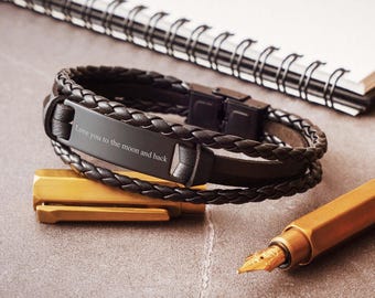 May include: A black braided leather bracelet featuring a rectangular metal plate engraved with the text Love you to the moon and back. The bracelet includes a black metal clasp and is positioned near a gold fountain pen and a spiral notebook.