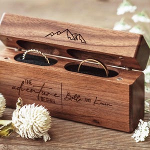 May include: A rectangular wooden ring box with a hinged lid featuring an engraved mountain graphic. The front is laser-engraved with the text "THE ADVENTURE BEGINS | Bella and Kevin". Inside are two black velvet slots holding a twisted gold ring and a plain gold band.