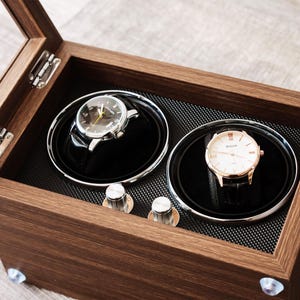 May include: A rectangular wooden watch winder box with a hinged glass lid, featuring two circular rotating compartments. Inside, two wristwatches with black leather straps are mounted on black cushions. Two silver control knobs are visible.