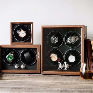 May include: A collection of three wooden watch winders with dark brown frames and black interiors. The largest unit features a glass front with a white letter M and the name Morgan printed on it, housing four watches. Two smaller units hold one and two watches.