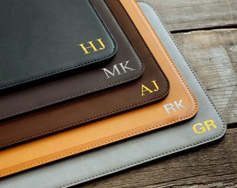 May include: A stack of five rectangular desk mats in black, dark brown, medium brown, tan, and gray. Each mat features stitched edges and a personalized two-letter monogram in gold or silver foil, specifically HJ, MK, AJ, RK, and GR.