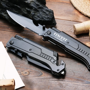 May include: Two matte black folding knives with serrated blades and integrated bottle openers. One knife is open, displaying the name VINCENT and the date 11.05.2024 engraved on the handle. The other knife is closed, showing a textured grip.